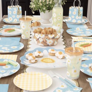 May include: A table setting with a blue and white daisy pattern on the plates, napkins, and cups. There are also yellow and white checkered napkins and a bowl of croissants.