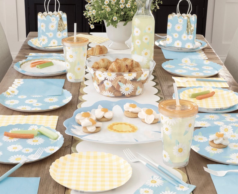 May include: A table setting with a blue and white daisy pattern on the plates, napkins, and cups. There are also yellow and white checkered napkins and a bowl of croissants.