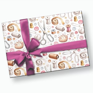 May include: A white gift box with a pink ribbon and bow. The box is wrapped in paper with a pattern of sewing supplies, including buttons, needles, thread, scissors, and measuring tape.