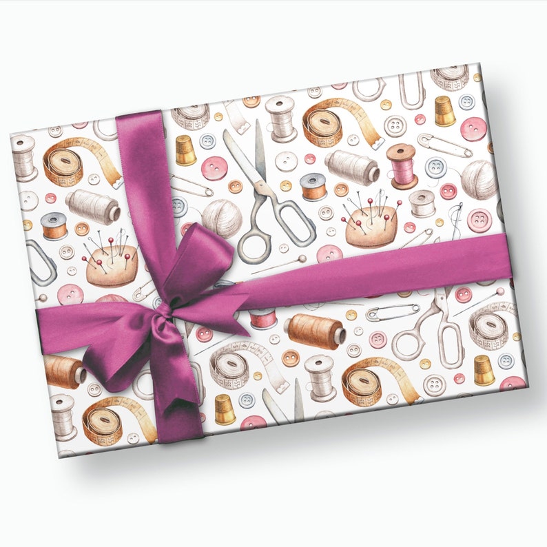 May include: A white gift box with a pink ribbon and bow. The box is wrapped in paper with a pattern of sewing supplies, including buttons, needles, thread, scissors, and measuring tape.