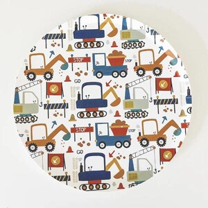 May include: A white, round plate with a repeating pattern of construction vehicles in blue, orange, and gray. The design includes excavators, dump trucks, and cranes, along with stop signs, arrows, and traffic cones. The plate has a diameter of approximately 10 inches.
