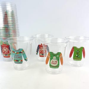 May include: Clear plastic cups with colorful Christmas sweater designs. The cups feature various sweater patterns in red, green, blue, and orange, with some including text like "Merry Christmas" and "Let it Snow!" A stack of cups is visible.