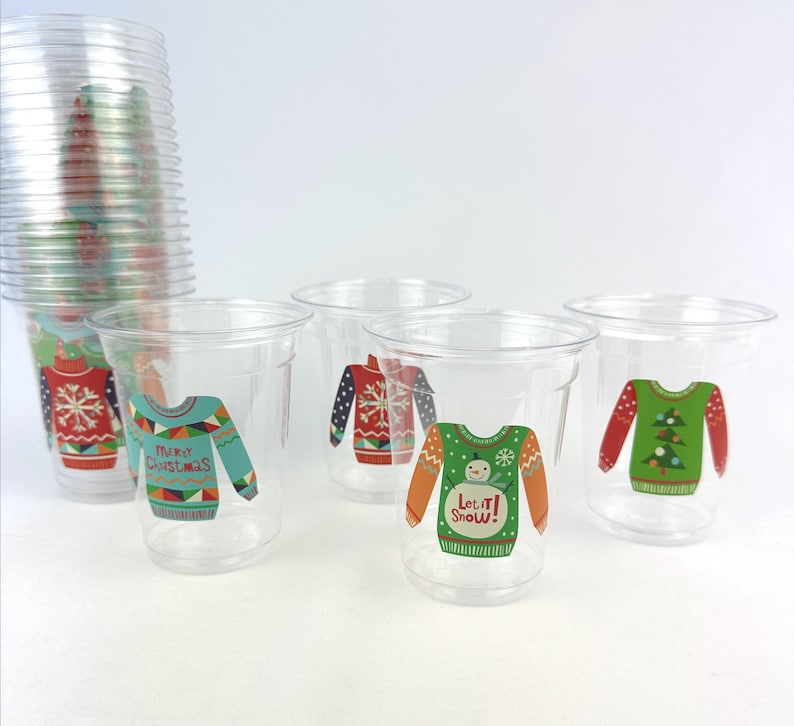 May include: Clear plastic cups with colorful Christmas sweater designs. The cups feature various sweater patterns in red, green, blue, and orange, with some including text like "Merry Christmas" and "Let it Snow!" A stack of cups is visible.