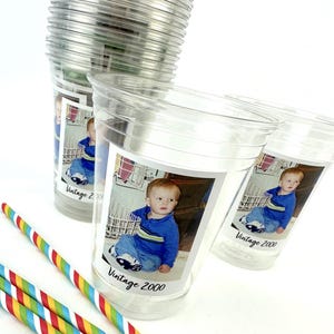 Puede incluir: Vasos de pl&aacute;stico transparente con una foto de beb&eacute; y el texto "Vintage 2000". Varias pajitas de rayas de colores est&aacute;n en primer plano. Los vasos est&aacute;n apilados sobre una superficie blanca.
