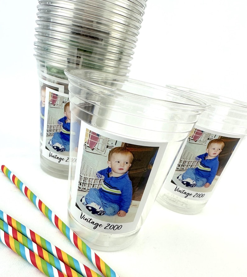 Puede incluir: Vasos de pl&aacute;stico transparente con una foto de beb&eacute; y el texto "Vintage 2000". Varias pajitas de rayas de colores est&aacute;n en primer plano. Los vasos est&aacute;n apilados sobre una superficie blanca.