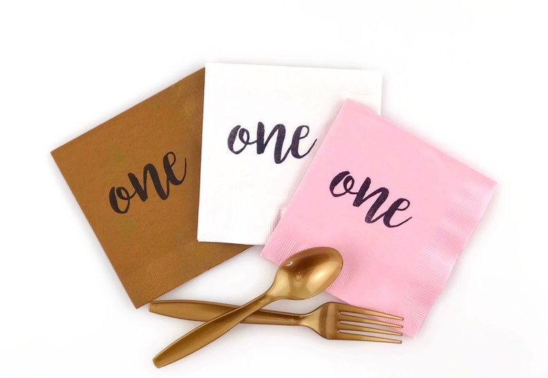 May include: Three paper napkins with the word "one" printed on them in black script. The napkins are brown, white, and pink. There are also two gold plastic utensils, a fork and a spoon.