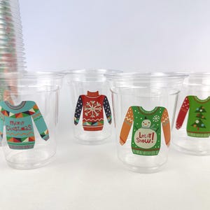 May include: Clear plastic cups with colorful Christmas sweater designs. The designs include a teal sweater with "Merry Christmas", a red sweater with a snowflake, and a green sweater with a snowman and "Let it Snow!" text.