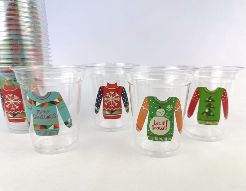 May include: Clear plastic cups with colorful Christmas sweater designs. The designs include a teal sweater with "Merry Christmas", a red sweater with a snowflake, and a green sweater with a snowman and "Let it Snow!" text.