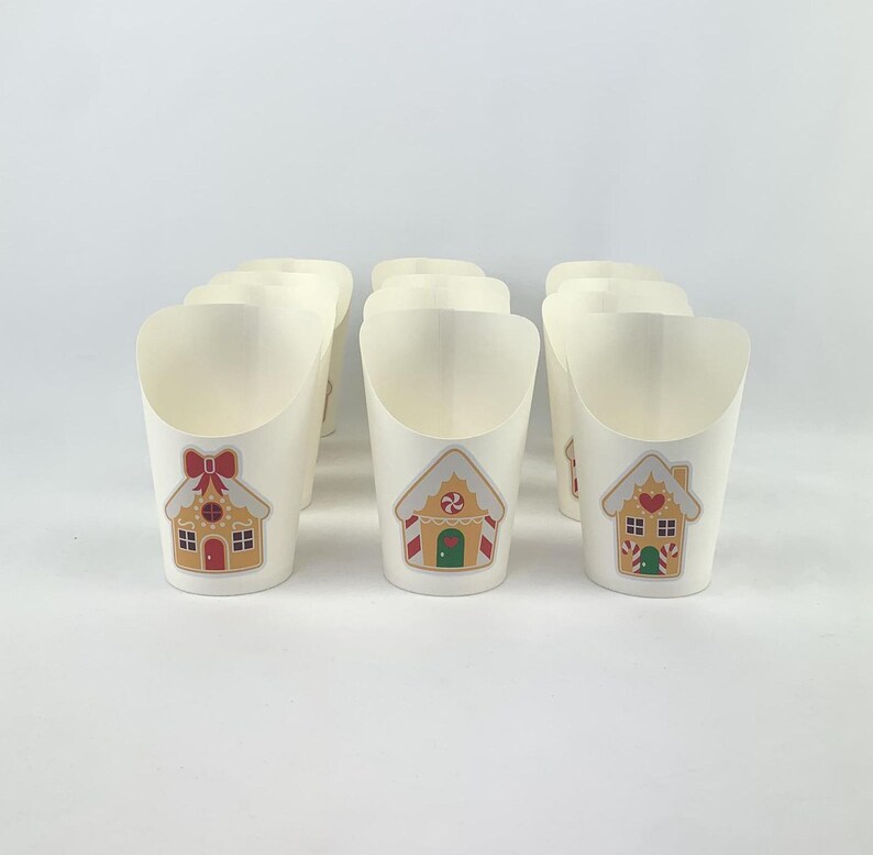 Puede incluir: Ocho vasos de papel blancos con un dise&ntilde;o de casa de jengibre. Las casas est&aacute;n decoradas con detalles rojos y verdes.
