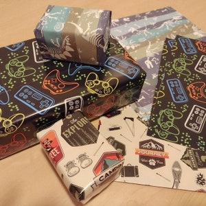 May include: A collection of gift wrap featuring various designs. One design features a black and white pattern of video game controllers and joysticks. Another design features a blue and white striped pattern with dinosaur skeletons. A third design features a white background with a variety of camping-themed illustrations and the text "The Explorer", "Camp", "Journey", and "Adventure".