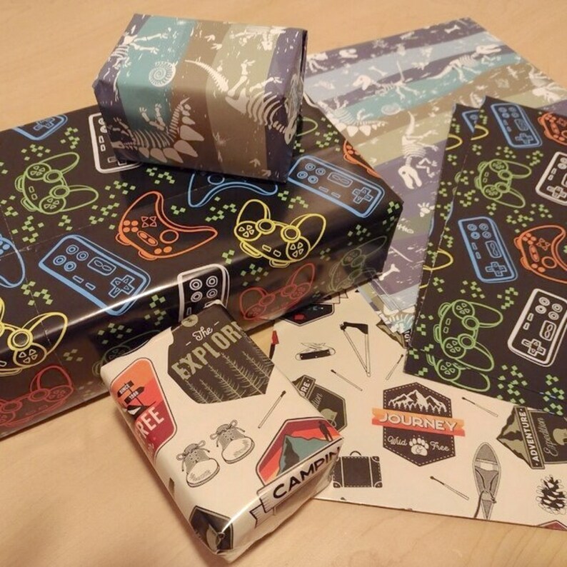 May include: A collection of gift wrap featuring various designs. One design features a black and white pattern of video game controllers and joysticks. Another design features a blue and white striped pattern with dinosaur skeletons. A third design features a white background with a variety of camping-themed illustrations and the text "The Explorer", "Camp", "Journey", and "Adventure".