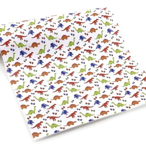 以下が含まれることがあります： 赤、緑、青、オレンジ色のカラフルな恐竜が繰り返し描かれた白い紙。恐竜は様々なポーズをしており、足跡がデザイン全体に散らばっています。紙は片方の端が丸まっています。