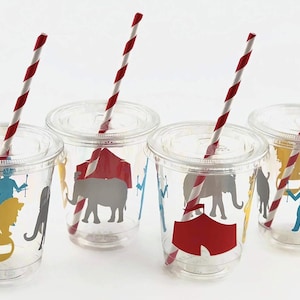 Puede incluir: Cuatro vasos de plástico transparente con pajitas de rayas rojas y blancas. Cada vaso tiene un diseño diferente con temática de circo, que incluye un elefante, un león, una tienda de campaña y un maestro de ceremonias.