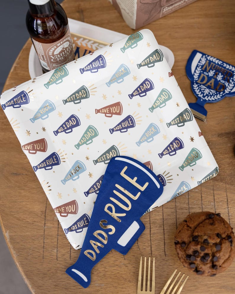 K&ouml;nnte beinhalten: Ein wei&szlig;er Pappteller mit einem goldenen und blauen Megaphon-Muster. Die Megaphone haben verschiedene Spr&uuml;che darauf, darunter "Dads Rule", "Best Dad", "You Rock", "Love You" und "No. 1 Dad". Es gibt einen blauen Megaphon-Ausschnitt mit dem Text "Dads Rule" darauf.