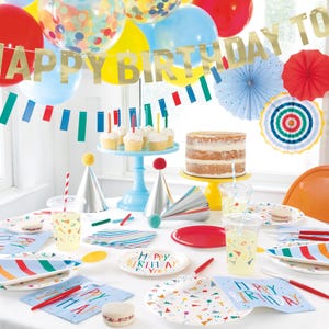 May include: A birthday party table setting with colorful paper plates, napkins, and cups. The table is decorated with a banner that says "Happy Birthday to", colorful balloons, and party hats. There is a cake on a blue stand and a red and white striped straw in a clear glass.