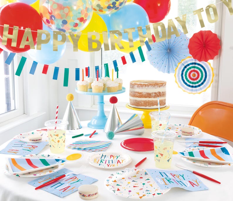 May include: A birthday party table setting with colorful paper plates, napkins, and cups. The table is decorated with a banner that says "Happy Birthday to", colorful balloons, and party hats. There is a cake on a blue stand and a red and white striped straw in a clear glass.
