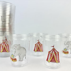 Puede incluir: Vasos de plástico transparente con ilustraciones de circo. Los vasos presentan imágenes de una carpa de circo roja y blanca y un elefante gris actuando sobre un tambor. Una pila de vasos es visible a la izquierda.