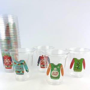 May include: Clear plastic cups with colorful Christmas sweater designs. The cups feature various sweater patterns with text that says "Merry Christmas" and "Let it Snow!" A stack of cups is visible in the background.