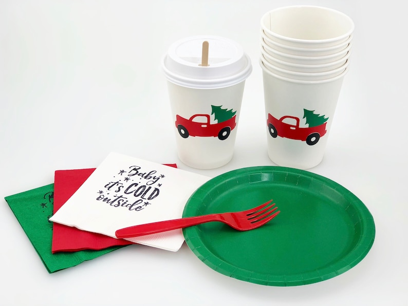 May include: A festive holiday party set featuring white paper cups with a red truck and Christmas tree design, green plates, red forks, and napkins with the text "Baby it's cold outside."