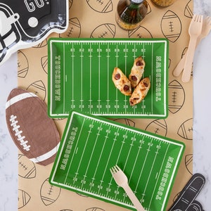 May include: Football-themed party supplies, including green field-shaped plates with "TOUCHDOWN" text, a football-shaped cutout, and a foam finger. A black helmet-shaped plate with "LET'S GO!" text is also present. The scene includes wooden forks and food.