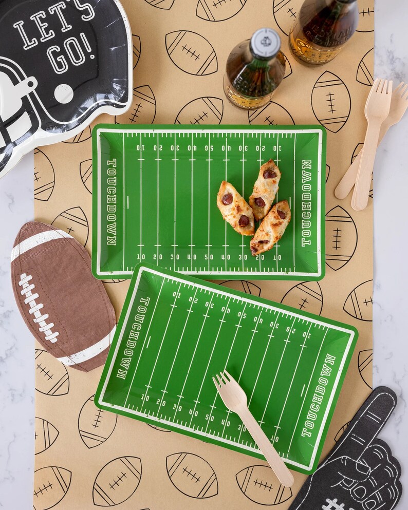 May include: Football-themed party supplies, including green field-shaped plates with "TOUCHDOWN" text, a football-shaped cutout, and a foam finger. A black helmet-shaped plate with "LET'S GO!" text is also present. The scene includes wooden forks and food.