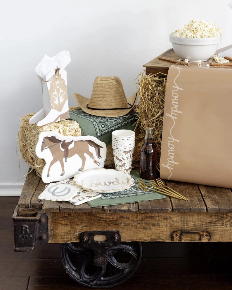 May include: A rustic wooden cart displays cowboy-themed party supplies. Items include a cowboy hat, horse-shaped plates, cups, napkins, and a large brown bag with "howdy" written on it. A bowl of popcorn and hay bales complete the scene.