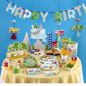 May include: A festive birthday party setup with a blue backdrop and a yellow tablecloth. The scene includes a cake, plates, cups, and decorations featuring a road and vehicle theme. A banner spells out "HAPPY BIRTH".