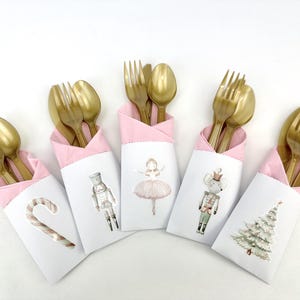 Op de afbeelding: Een set van vijf witte en roze bestekhouders met gouden vorken en lepels. Elke houder heeft een feestelijke illustratie: een zuurstok, een notenkraker, een ballerina, een muis en een kerstboom. Perfect voor de feestdagen.