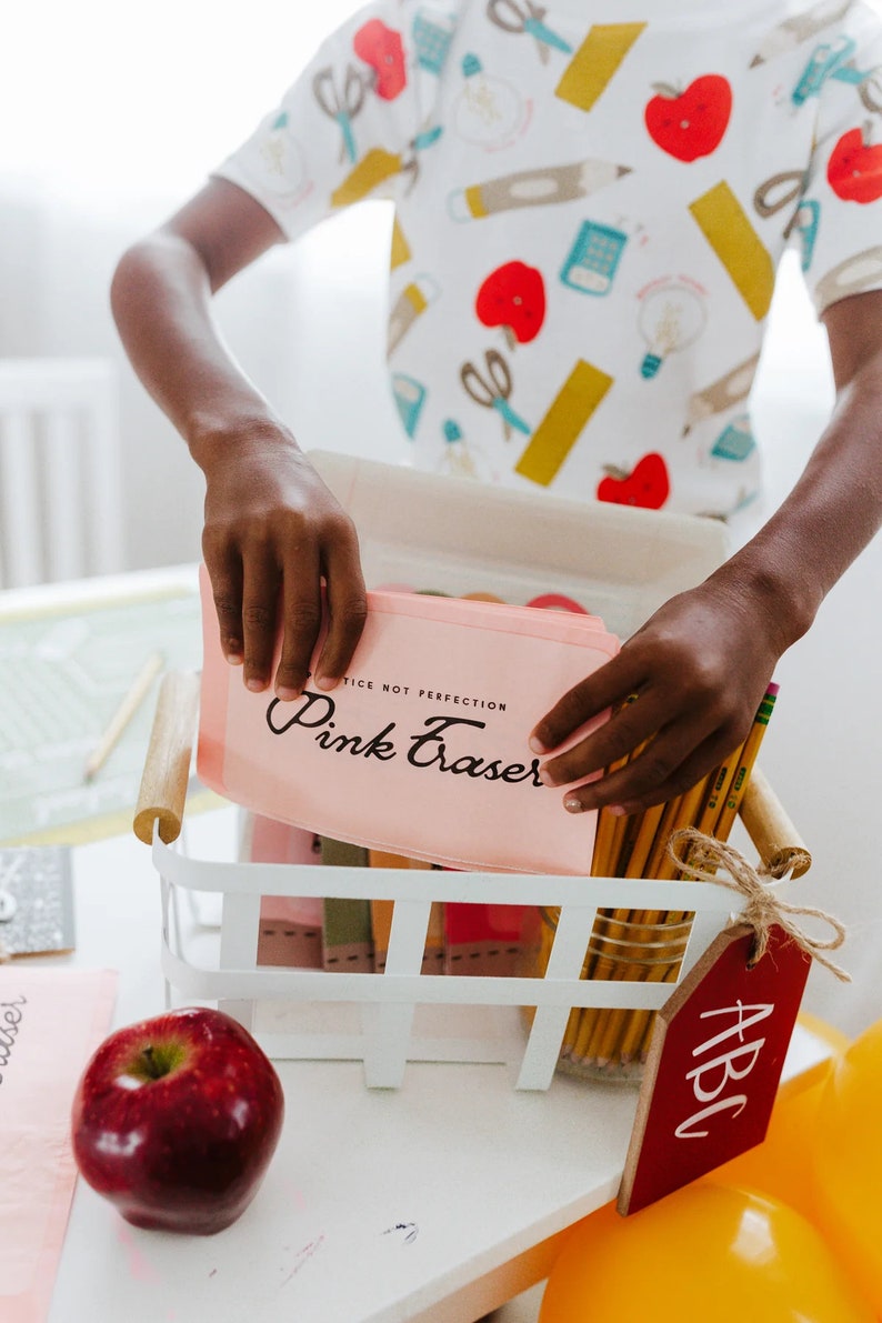 May include: A white basket filled with pencils and pink erasers. The pink eraser case has the words "Pink Eraser" printed on it. A red apple and a wooden tag with "ABC" are also visible. The person is wearing a white shirt with school-themed graphics.