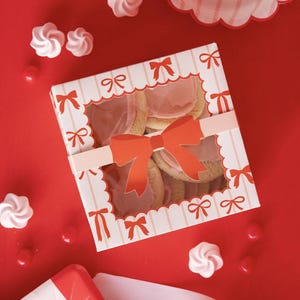 May include: A gift box of cookies, decorated with red bows and a large central bow. The box has a window showing the cookies inside. The image is set against a red background with sweets and meringues.