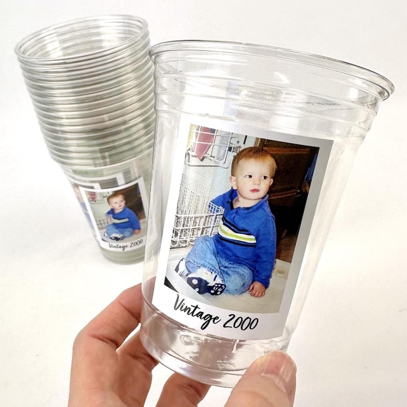 Puede incluir: Vasos de pl&aacute;stico transparente con una foto de un ni&ntilde;o peque&ntilde;o. La foto est&aacute; etiquetada como "Vintage 2000". El ni&ntilde;o lleva una camisa azul y vaqueros. En el fondo se ve una pila de vasos similares.