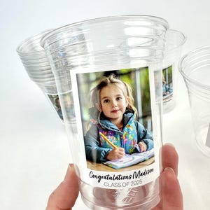 May include: Clear plastic cups with a printed label featuring a young girl. The label reads "Congratulations Madison CLASS OF 2025". The girl is wearing a colourful jacket and holding a pencil.