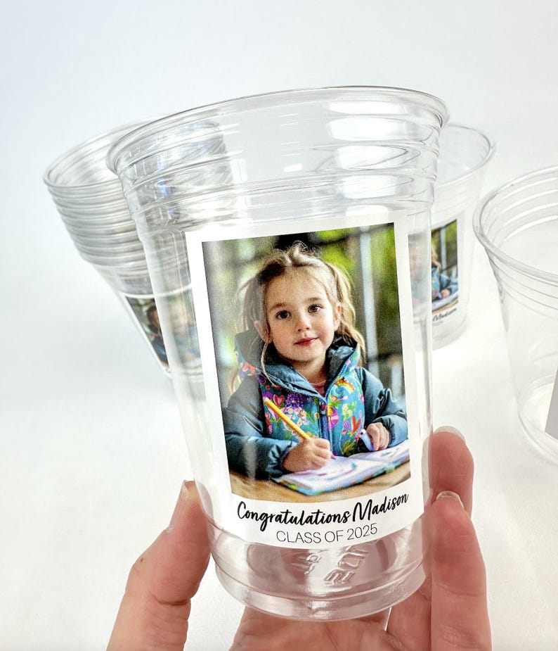 May include: Clear plastic cups with a printed label featuring a young girl. The label reads "Congratulations Madison CLASS OF 2025". The girl is wearing a colorful jacket and holding a pencil.