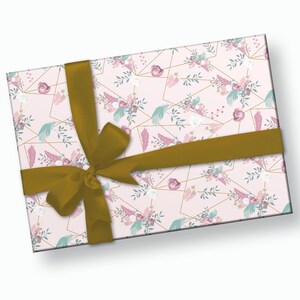 K&ouml;nnte beinhalten: Ein rechteckiger Geschenkkarton, der in rosa floral gemustertes Papier mit goldenen geometrischen Linien verpackt ist. Das Design umfasst rosa Rosen, wei&szlig;e Blumen und t&uuml;rkisfarbene Federn. Ein goldenes Satinband ist um die Schachtel gebunden.