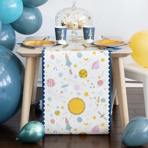 May include: A white table runner with a blue scalloped border features a space-themed design with planets, stars, rockets, and the text "Blast Off!" The runner is perfect for a space-themed party.