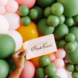 May include: A hand holds a pink card with the words "Pink Eraser" and "Practice Not Perfection" in black script. The card is set against a backdrop of green, pink, white, and yellow balloons, creating a colorful and festive scene.