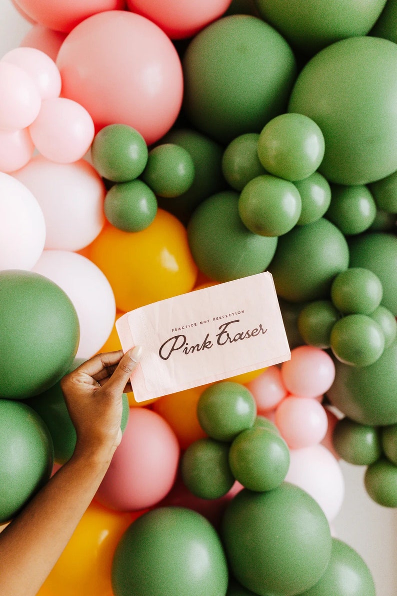 May include: A hand holds a pink card with the words "Pink Eraser" and "Practice Not Perfection" in black script. The card is set against a backdrop of green, pink, white, and yellow balloons, creating a colorful and festive scene.
