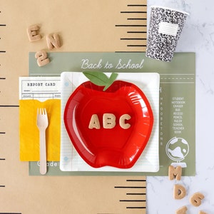 May include: A festive back-to-school arrangement featuring a red apple-shaped plate with ABC letters, a paper cup with a black and white speckled design, and a yellow napkin with a wooden fork. The words "Back to School" are visible.