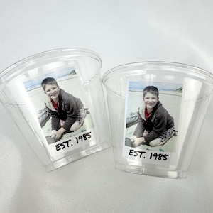 Puede incluir: Dos vasos de plástico transparente con una foto de un niño sonriendo. La imagen es de un niño en una playa. El texto "EST. 1985" está impreso en una etiqueta blanca debajo.