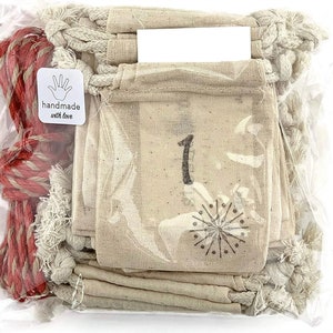 Puede incluir: Una bolsa de pl&aacute;stico transparente contiene peque&ntilde;as bolsas de tela beige con el n&uacute;mero "1" impreso. Un dise&ntilde;o de copo de nieve est&aacute; debajo del n&uacute;mero. Tambi&eacute;n se ven una cuerda roja y blanca y una pegatina "handmade with love".