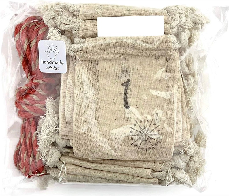 Puede incluir: Una bolsa de pl&aacute;stico transparente contiene peque&ntilde;as bolsas de tela beige con el n&uacute;mero "1" impreso. Un dise&ntilde;o de copo de nieve est&aacute; debajo del n&uacute;mero. Tambi&eacute;n se ven una cuerda roja y blanca y una pegatina "handmade with love".