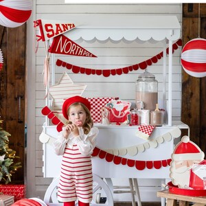 May include: A white wooden cart decorated with red and white paper garland, red and white paper lanterns, and a red and white Santa banner. A young girl wearing a red and white striped dress and a red beret is standing in front of the cart. The cart is decorated with a red and white checked tablecloth and a red and white Santa plate.