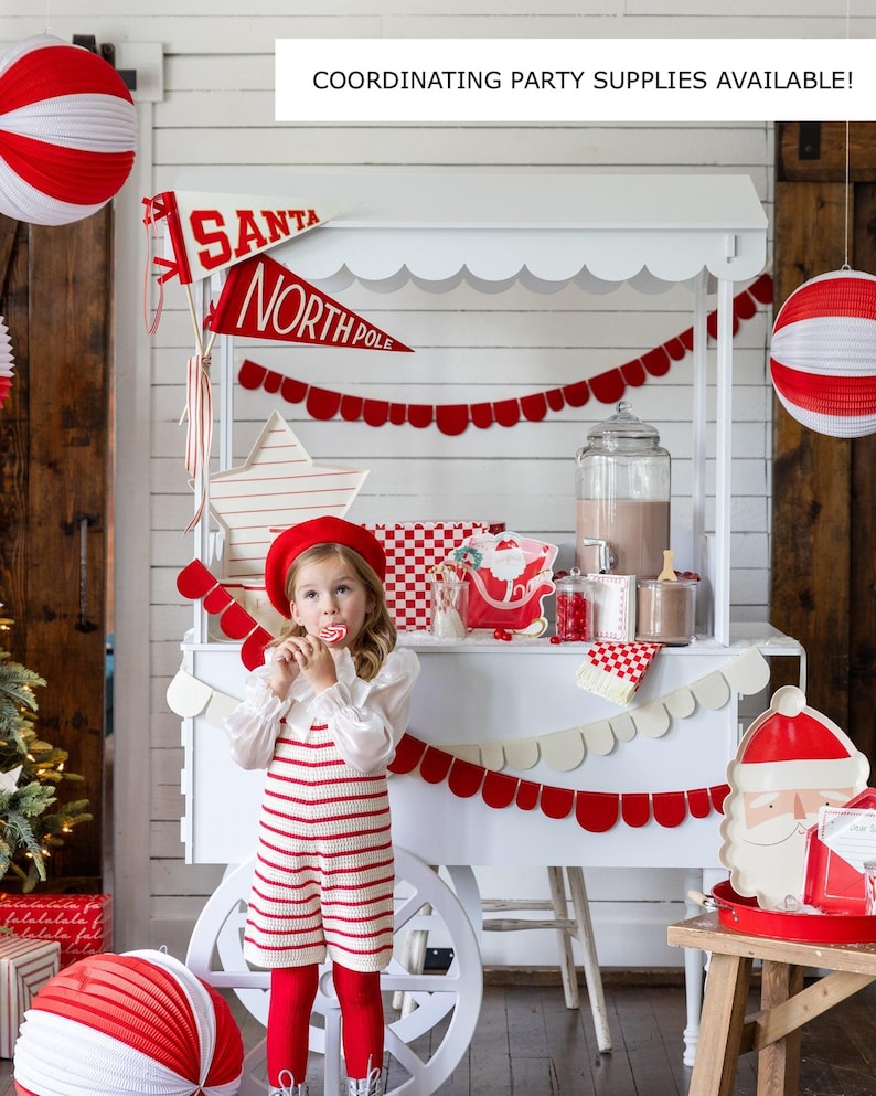May include: A white wooden cart decorated with red and white paper garland, red and white paper lanterns, and a red and white Santa banner. A young girl wearing a red and white striped dress and a red beret is standing in front of the cart. The cart is decorated with a red and white checked tablecloth and a red and white Santa plate.