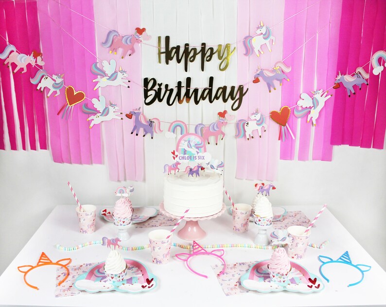 May include: A unicorn-themed birthday party table setting with pink and white paper goods, a unicorn cake with a rainbow topper, and a unicorn headband. The cake says "Chloe is Six".