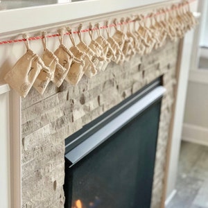 Puede incluir: Calendario de Adviento con peque&ntilde;as bolsas de tela beige colgadas de una cuerda roja y blanca. Cada bolsa est&aacute; numerada y decorada con un dise&ntilde;o sencillo. El calendario se muestra sobre una chimenea con fachada de piedra.