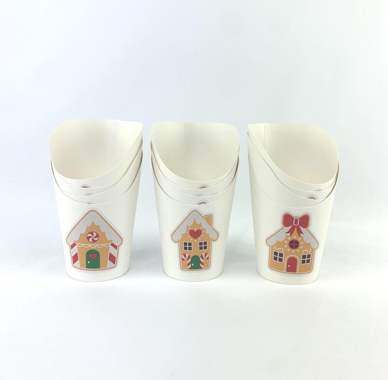 Puede incluir: Tres vasos de papel blancos con dise&ntilde;os de casas de jengibre. Cada vaso tiene un dise&ntilde;o diferente con una casa de jengibre con detalles de caramelo. Los vasos est&aacute;n apilados uno encima del otro.