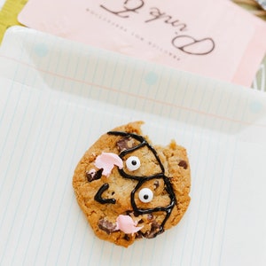 May include: A chocolate chip cookie decorated with black icing, pink frosting, and two edible white eyes. The cookie sits on a white plate with blue lines. A pink card with the text "Pink Roses" is in the background.