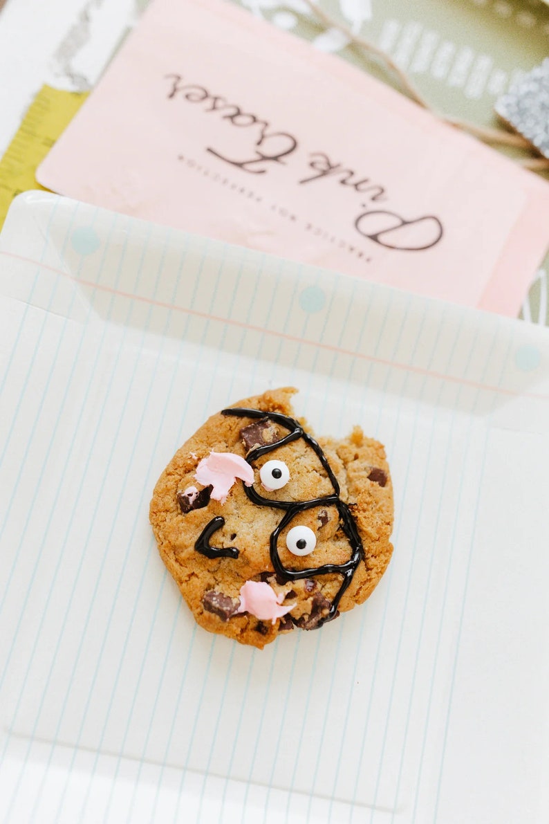May include: A chocolate chip cookie decorated with black icing, pink frosting, and two edible white eyes. The cookie sits on a white plate with blue lines. A pink card with the text "Pink Roses" is in the background.