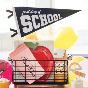 May include: A black wire basket filled with school-themed decorations. Includes a pennant that reads "first day of SCHOOL", a red apple-shaped plate, pink cards, and cupcake toppers. Yellow balloons are in the background.