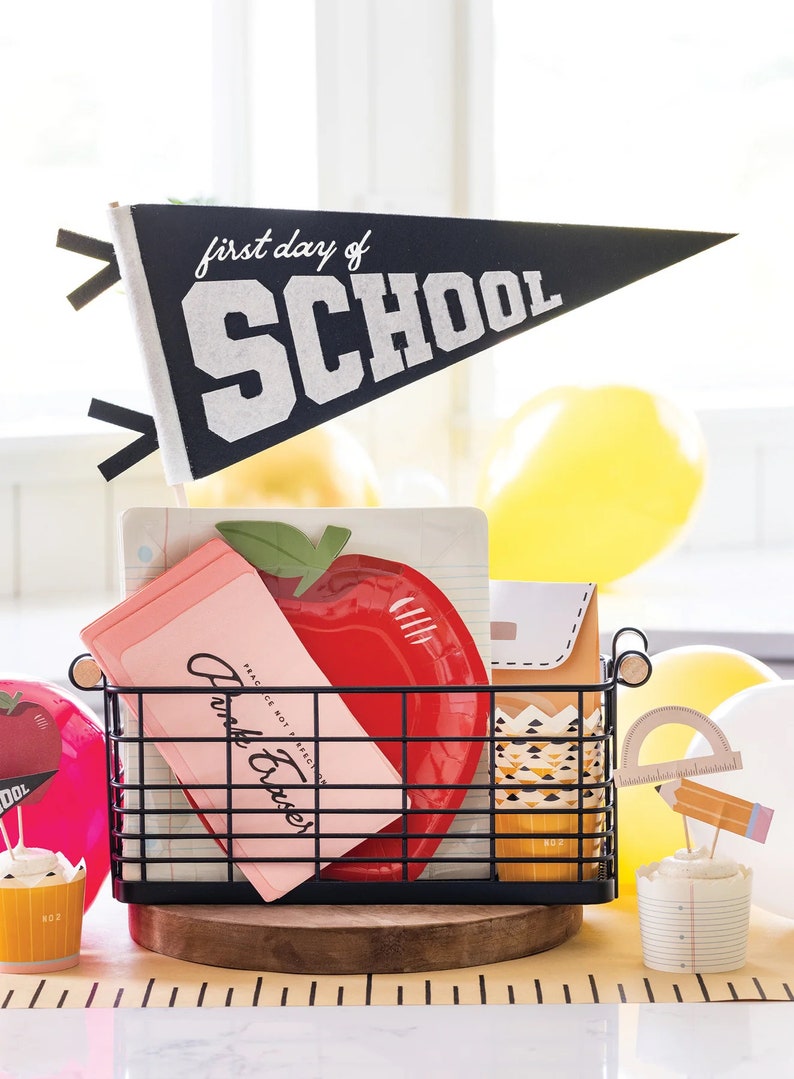 May include: A black wire basket filled with school-themed decorations. Includes a pennant that reads "first day of SCHOOL", a red apple-shaped plate, pink cards, and cupcake toppers. Yellow balloons are in the background.